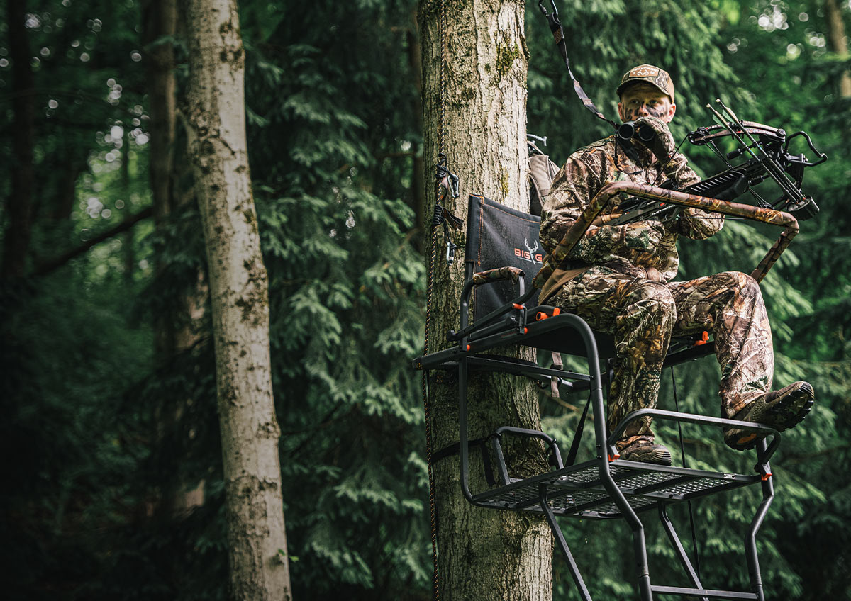 A hunter sitting in a ladder stand looking up from his binoculars.
