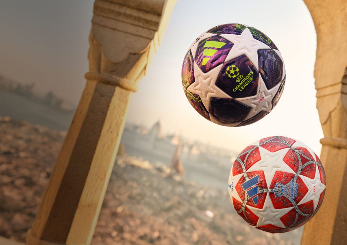 Two soccer balls mid-air. European city in the background.