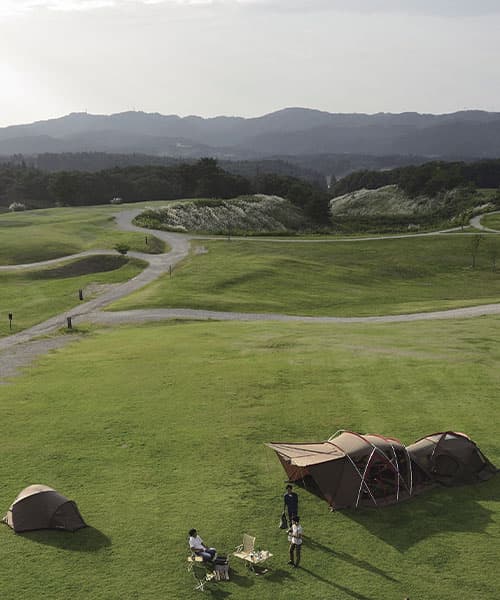 Campers standing in a green field next to their tent.