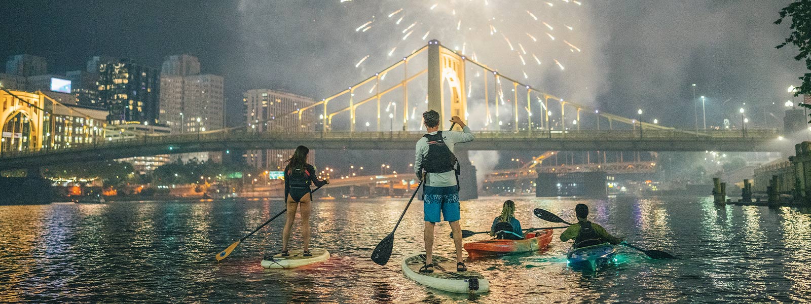 people stand-up paddle boarding in front of yellow Pittsburgh bridge and fireworks
