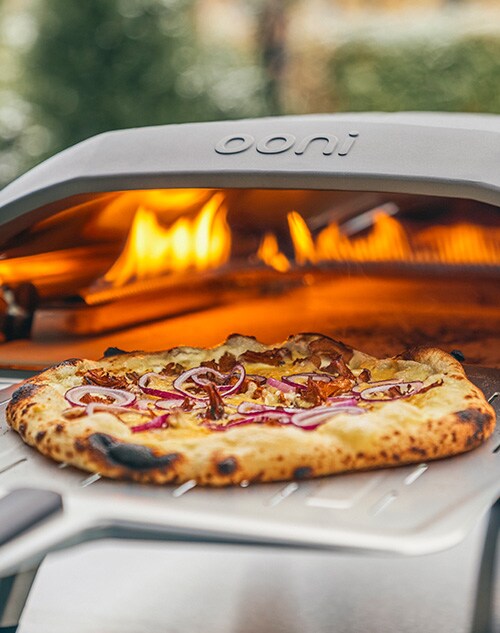 A pizza being taken out of an Ooni outdoor oven.