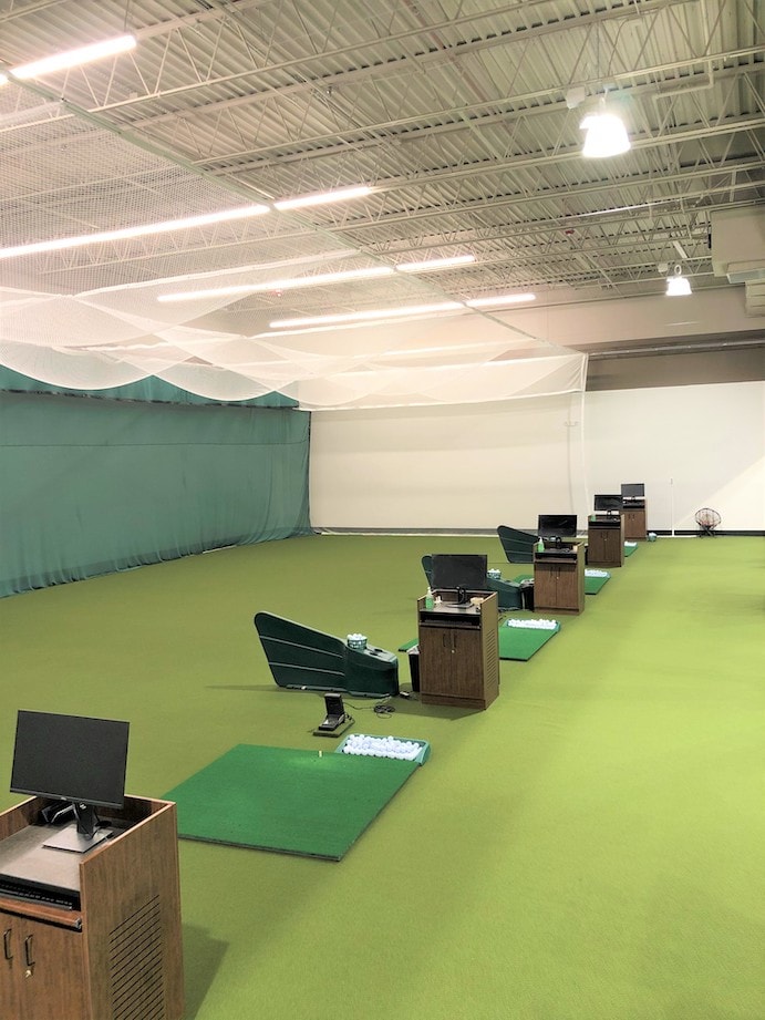 An open room with four range driving bays. Each one has a green tee pad, a container of golf balls, and a podium with a computer monitor. About 30 feet in front of the tee pads is a large green curtain.